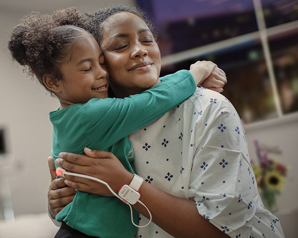 Dark skinned patient wearing hospital gown monitored by Masimo tetherless sensor hugging child.